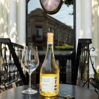 A bottle of white wine and an empty wine glass on a dark table in front of a white building with a large arched window and a hanging clock.